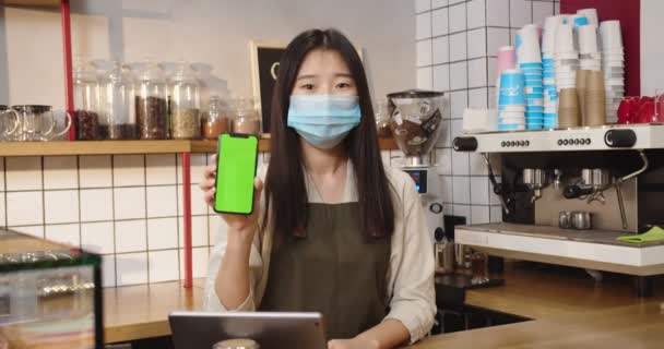 Attraktiv asiatisk kvinde iført medicinsk maske, stående bag bar og viser grøn skærm. Ung dame barista holder digital smartphone med chroma nøgle i cafe, coffeehouse. Lodret position. – Stock-video