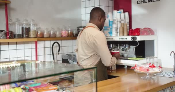 Retrato de jovem garçom masculino segurando xícara de café e sorrindo no café urbano. Bonito barista afro-americano a fazer bebidas e a entregar à câmara. Pequenas empresas, conceito de serviço. — Vídeo de Stock