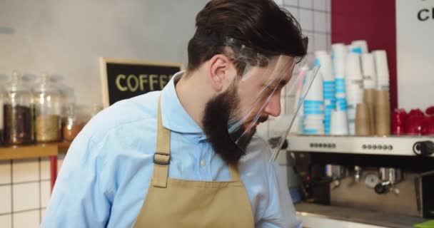 Retrato do proprietário do café macho alegre na limpeza da viseira do protetor facial, limpando a superfície da barra. Jovem homem caucasiano mesa de higienização, olhando para a câmera, cruzando os braços e sorrindo. Vírus, medidas de segurança. — Vídeo de Stock