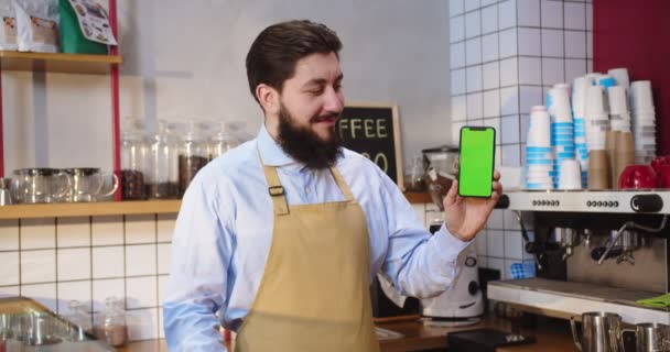 Barista masculino alegre segurando smartphone com tela verde no café. Jovem caucasiano olhando para a câmera, sorrindo e mostrando a chave cromática, posição vertical. Conceito de publicidade. — Vídeo de Stock