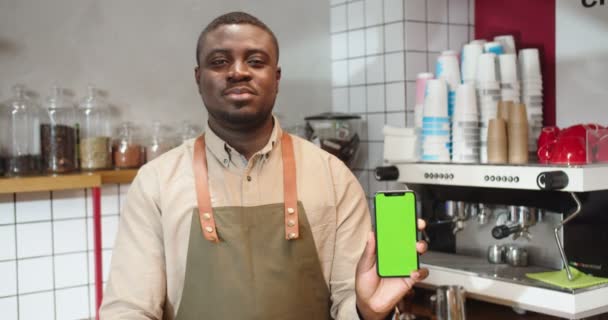 Barista masculino alegre segurando smartphone com tela verde no café. Adulto afro-americano olhando para a câmera, sorrindo e mostrando croma chave, posição vertical. Conceito de negócio. — Vídeo de Stock