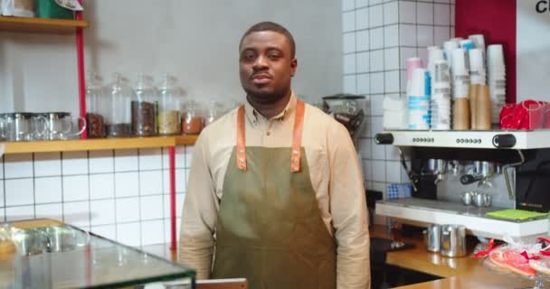 Varón barista guapo usando delantal y de pie en el mostrador en la cafetería. Jóvenes afroamericanos trabajando en una cafetería, mirando a la cámara y cruzando brazos. Pequeña empresa, concepto de compra. — Vídeo de stock
