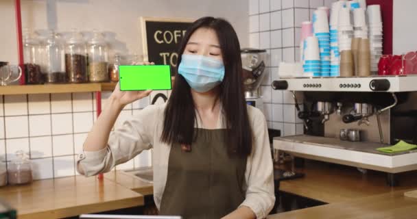 バーの後ろに立って緑色の画面でスマートフォンを保持エプロンで魅力的なアジアの女性。マスクの若い女性バリスタカメラを見て、笑顔とクロマキーを表示します。水平位置. — ストック動画