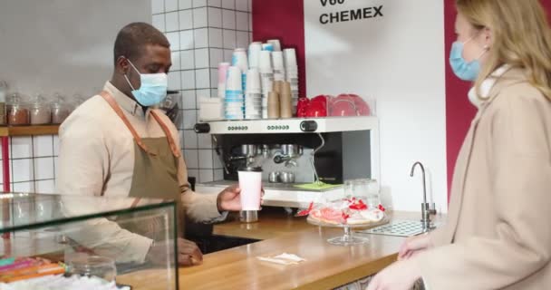 Zelfverzekerde mannelijke barista in medische beschermende masker het maken van koffie, geven aan vrouwelijke klant en afscheid nemen. Mooie blanke vrouw die een borrel meeneemt. Quarantaine, handel, virus. — Stockvideo