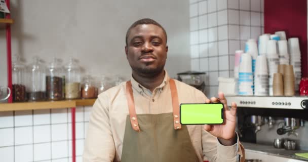 Bonito afro-americano cara de pé atrás da barra e segurando smartphone com chave chroma. Homem barista olhando para a câmera e mostrando tela verde no café. Posição vertical. Negócios. — Vídeo de Stock