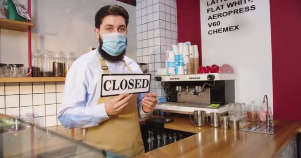 Dissapointed young man in protective medical mask standing behind bar. Caucasian male barista turning sign from OPEN to CLOSED because of lockdown, quarantine. Crisis, business, pandemic concept. — Stock Video