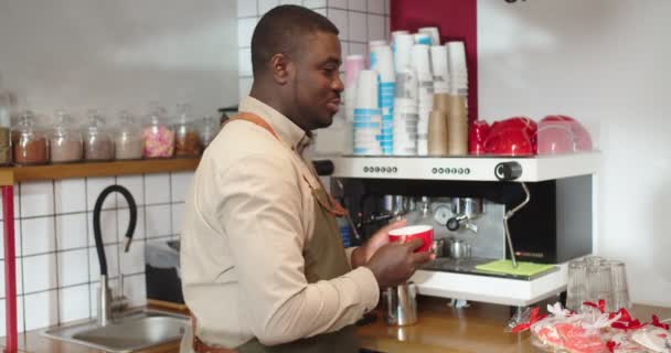 コーヒーマシンで作ったドリンクを飲みながらカフェで働くアフリカ系アメリカ人の陽気な男。大人の男性バリスタカメラを見て、笑顔とカップを保持。ビジネス、コーヒーハウスのコンセプト. — ストック動画