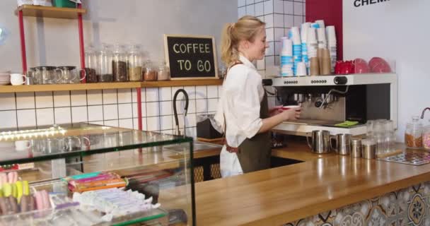 Симпатичная бариста работает в современной кофейне, делает напитки на кофеварке. Молодая белая женщина пьет чашку, ставит бар, смотрит в камеру и улыбается. Бизнес. — стоковое видео