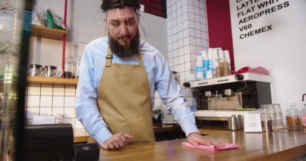 Homem caucasiano confiante na viseira do escudo facial a desinfectar a mesa do bar no café. Homem adulto barista limpando e limpando seu local de trabalho no café. Vírus, medidas de segurança, conceito de quarentena. — Vídeo de Stock