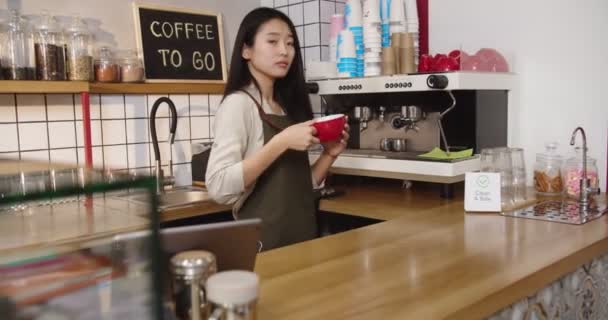 Krásná žena barista amking kávu, přičemž šálek a dát ho na barový stůl v kavárně. Mladá Asiatka drží nápoj, dívá se do kamery a usmívá se. Čistý sejf. Služba, práce na částečný úvazek. — Stock video