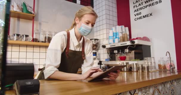 Succesvolle vrouwelijke koffiehuis eigenaar leunend op de bar en werken op tablet. Volwassen blanke vrouw in medisch masker typen en het aannemen van orders op gadget in cafe. Technologieën, bedrijfsconcept. — Stockvideo