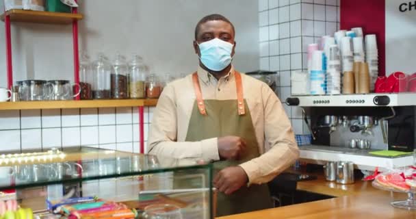 Porträtt av charmiga manliga barista bär förkläde och arbetar i små kafé. Ung afroamerikansk kille i skyddande medicinsk mask tittar på kameran och korsar armarna. Pandemi, karantän. — Stockvideo