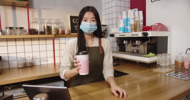 POV Barista femenina feliz dando taza de bebida al cliente y sonriendo. Jóvenes camareros asiáticos en máscara médica acechando orden y diciendo adiós. Bebiendo a mano. Servicio, concepto de cuarentena. — Vídeos de Stock