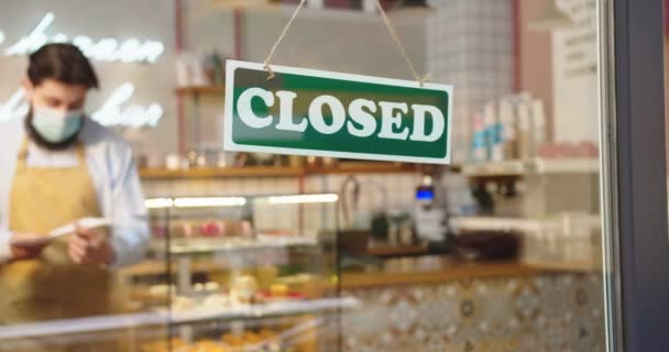 Außenansicht eines männlichen Baristas, der die vorübergehende Schließung des Kaffeehauses aufgrund von COVID-19 ankündigt. Erwachsener Mann blättert Schild OPEN, legt Papier auf Sorry, wir sind geschlossen. Coronavirus, Wirtschaft. — Stockvideo