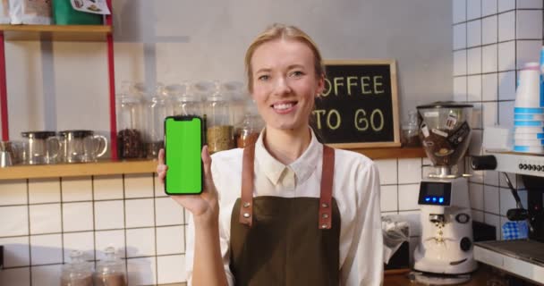 Πορτρέτο του γοητευτικό Καυκάσιος γυναίκα στέκεται πίσω από μπαρ και τρύπα smartphone με chroma κλειδί. Νεαρή μπαρίστα που κοιτάζει την κάμερα, χαμογελώντας και δείχνοντας πράσινη οθόνη στην καφετέρια. Έννοια εργασίας. — Αρχείο Βίντεο