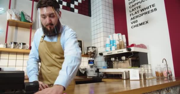 Snygg ung man städa och sanera sin arbetsplats i caféet. Vit manlig barista i ansiktsskydd visir torka ner bar, tittar på kamera och korsning armar. Karantänkoncept. — Stockvideo