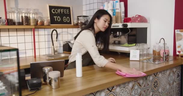 Çalışkan bayan barmen kafeteryada barmenlik yapıyor. Yetişkin Asyalı kadın dezenfekte masaları dezenfekte ediyor, kameraya bakıyor ve kafede gülümsüyor. Virüs, mikrop konsepti. — Stok video