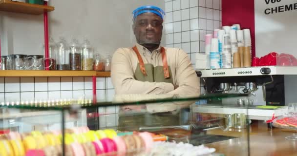 Profesyonel erkek barista önlük giyiyor ve karantina sırasında kahve dükkanında çalışıyor. Koruyucu yüz kalkanı takmış genç Afro Amerikalı adam kameraya bakıyor ve kollarını kavuşturuyor. Salgın. — Stok video