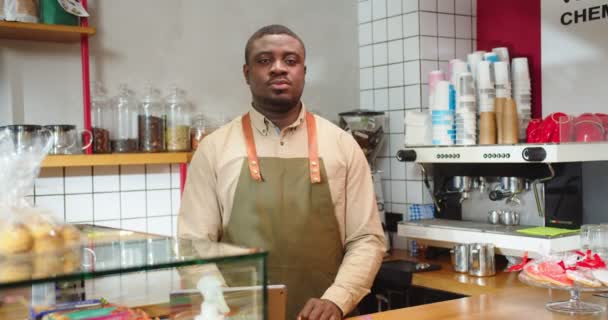 Modern kahve dükkanında minimalist iç mimarisi olan profesyonel bir baristanın portresi. Genç Afro-Amerikalı bir adam, barın önünde durmuş kameraya bakıyor ve kollarını kavuşturuyor. Kafe, iş konsepti. — Stok video