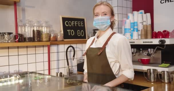 Retrato de una barista profesional detrás de una mesa de bar en una cafetería. Mujer caucásica adulta en máscara médica protectora mirando a la cámara y sonriendo. Virus, pandemia, concepto de cuarentena. — Vídeos de Stock