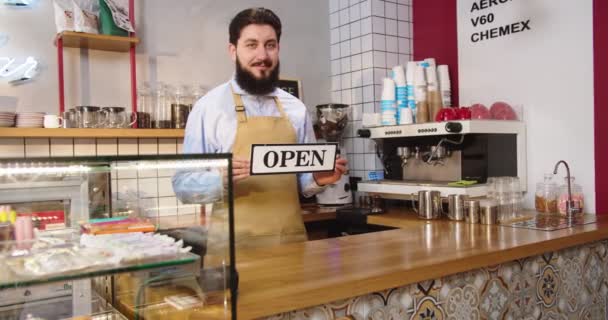 Hermoso empleado de café caucásico de pie detrás de la barra y mostrando el signo ABIERTO. Joven barista de uniforme de delantal mirando a la cámara y sonriendo en la cafetería. Negocios, concepto de start-up. — Vídeos de Stock