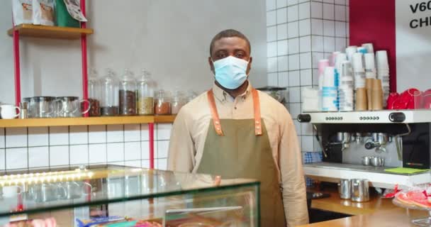 Retrato del apuesto dueño de una pequeña empresa parado detrás de la barra en la cafetería. Hombre afroamericano adulto con máscara médica protectora con delantal mirando a la cámara en la tienda. Concepto de servicio. — Vídeos de Stock