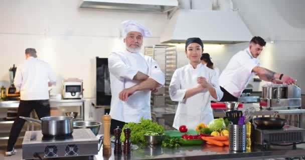 Happy senior Kaukasische mannelijke chef-kok staan in restaurant keuken op het werk met jonge Aziatische vrouwelijke kok kijken naar de camera en glimlachen. Mensen die werken op de achtergrond, koken concept — Stockvideo