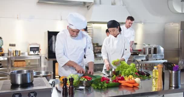 Stary biały mężczyzna kroi pomidory, podczas gdy azjatycka młoda kobieta gotuje pranie sałatki pracując w restauracji. Restauracja kuchnia, krojenie warzyw, przygotowywanie posiłków. Gotowanie, koncepcja kulinarna — Wideo stockowe