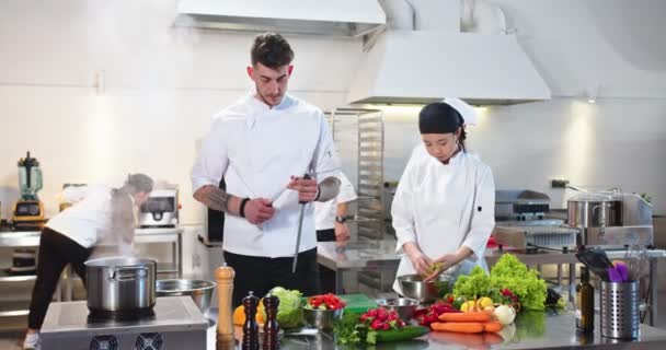 Team misto di cuochi che preparano il cibo lavorando nel ristorante. Il giovane chef caucasico affila il coltello. Asiatico giovane bella donna lavaggio insalata. Cucina del ristorante, preparazione del pasto. Concetto Cook — Video Stock