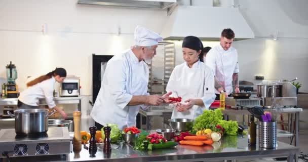 Słynny biały stary kucharz mówi i uczy swoją azjatycką młodą uczennicę przygotować specjalne danie w nowoczesnej kuchni pięciogwiazdkowej restauracji. Przebieg pracy, proces pracy, branża kulinarna — Wideo stockowe