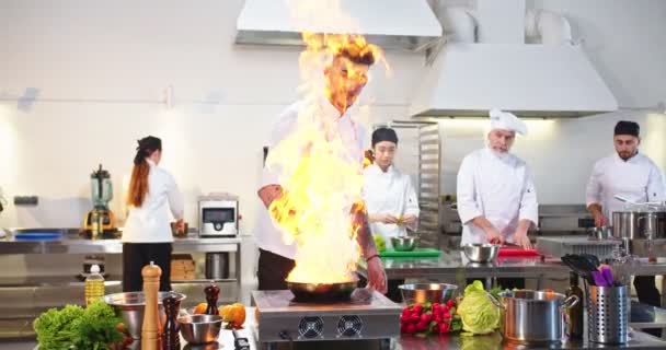 손에 익은 젊은 백인 남자 요리사가 유명 한 식당 식당의 부엌에 갖다 버리는 재료들을 볶는 불을 가지고 워크를 만드는 일을 한다. 배경 작업을 하는 사람들. 요리하는 일 — 비디오