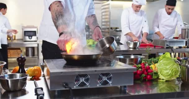 Primo piano di giovane esperto famoso chef maschio caucasico che lavora preparando pasto wok con fiamme arrostimento gettando ingredienti alla cucina del ristorante. Le persone di razza mista lavorano sullo sfondo. Concetto Cook — Video Stock