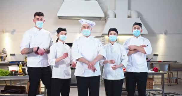Equipe de chefs multiétnicos e profissionais diversificados em máscaras médicas em pé na cozinha do restaurante e olhando para a câmera antes do dia útil durante a pandemia vívida. Culinária, conceito de trabalho — Vídeo de Stock