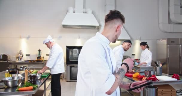Porträtt av ung vacker vit manlig kock står i restaurang kök skriva på surfplatta enhet och prata med upptagen kille kock arbetare visar ordning måltid. Arbetsflöde process, team som förbereder mat — Stockvideo