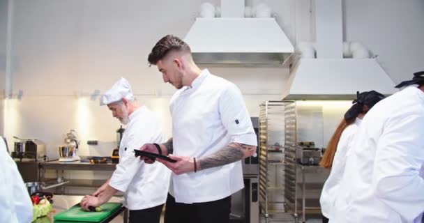 Mladý pohledný kavkazský kuchař kráčí v kuchyni restaurace a píše na tabletu zařízení, které kontroluje pracovní postupy a mluví s vytíženým kuchařem. skupina slavné přípravy jídla — Stock video