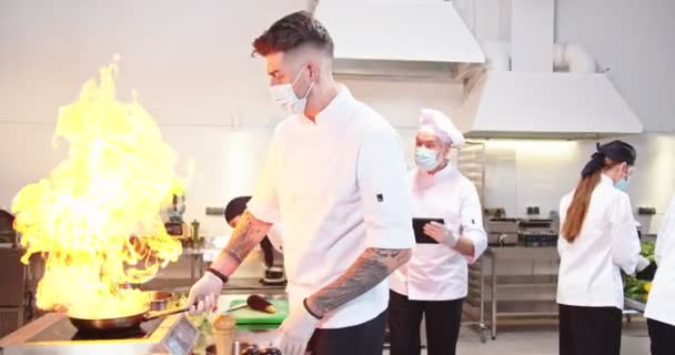 Caucasiano jovem cozinheiro do sexo masculino fazendo wok refeição com chamas, chefe de chef profissional homem sênior andando na cozinha do restaurante navegando no tablet supervisionando o controle do fluxo de trabalho de culinária Preparando alimentos, covid — Vídeo de Stock