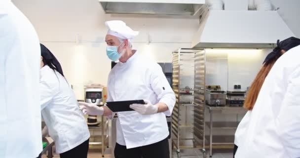 Καυκάσιος ανώτερος γέρος επαγγελματίας σεφ αφεντικό στην ιατρική μάσκα περπάτημα στην κουζίνα εστιατόριο πατώντας στη συσκευή tablet εποπτεία της διαδικασίας μαγειρικών εργασιών και της ροής εργασίας. Προετοιμασία των τροφίμων, covid καραντίνα — Αρχείο Βίντεο