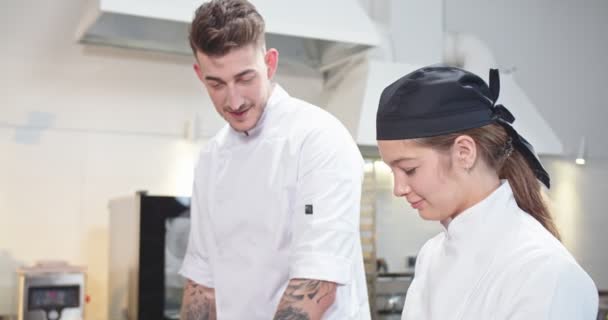 Fecha. Vista lateral do jovem jovem e alegre chef taching para preparar a refeição na cozinha do restaurante e falar com o colega de cozinha caucasiano feminino. Processo de trabalho, conceito culinário — Vídeo de Stock