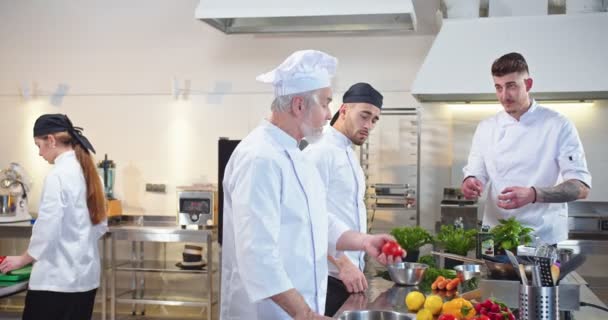 Caucasian male young famous chef speaking with cooks apprentices during working process at restaurant kitchen telling teaching them cooking. Team of chefs preparing food dish. Culinary concept — Stock Video