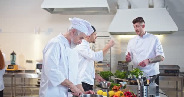 Hombre caucásico joven jefe gerente hablando con cocineros profesionales durante el proceso de trabajo en la cocina del restaurante diciendo plato ordenado. Equipo de chefs preparando comida. Concepto culinario — Vídeos de Stock