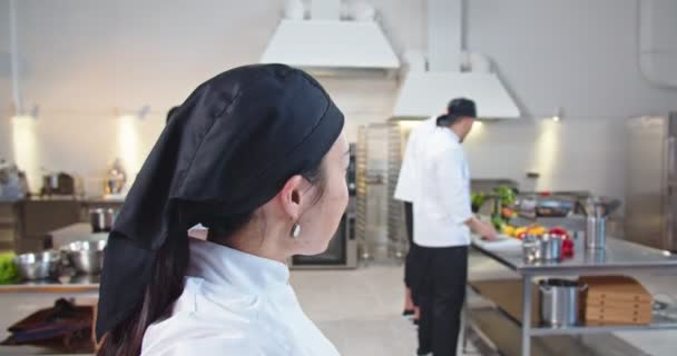 Närbild av asiatisk glad professionell kvinnlig kock videochatting stående i köket visar hennes team av kockar under arbetsprocessen POV av glada ung kvinna talar på videosamtal på restaurang online — Stockvideo