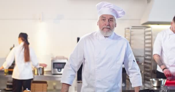 Primer plano retrato de alegre positivo pensionista hombre chef en sombrero mirando a la cámara de buen humor sonriendo mientras está de pie en el trabajo en la cocina del restaurante. Flujo de trabajo sobre fondo, concepto de cocina — Vídeos de Stock