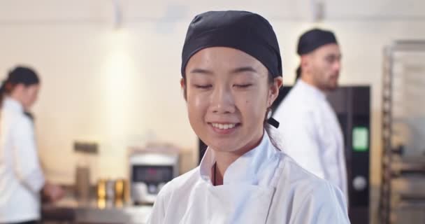 Close up retrato de alegre jovem asiático bela chef feminino em chapéu olhando para a câmera de bom humor sorrindo enquanto estava de pé no trabalho na cozinha do restaurante. Fluxo de trabalho em segundo plano, conceito de cozinha — Vídeo de Stock