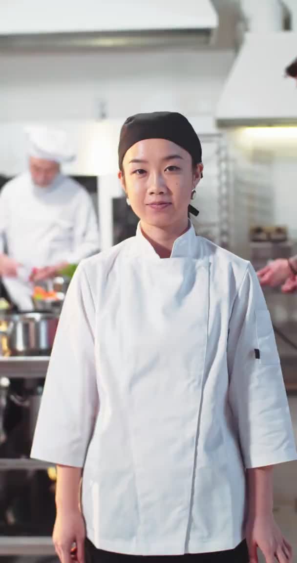 Verticale video van jonge vrolijke Aziatische professionele chef-kok kijkend naar camera staande in restaurant op het werk. Portret van een vrolijke vrouwelijke kok in de keuken. Positieve emoties, business concept van de voedingsindustrie — Stockvideo