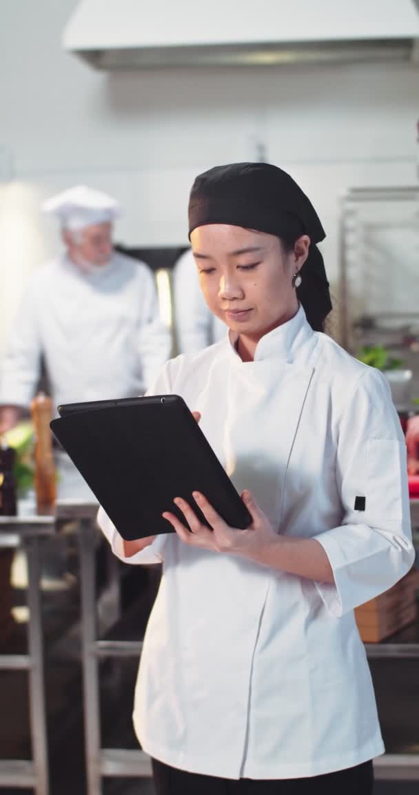 Porträtt av asiatiska vackra unga kvinna kock stående i restaurang kök i uniform knacka på surfplatta enhet surfa online på arbetsplatsen. Vertikala bilder. Begreppet kokyrke — Stockvideo