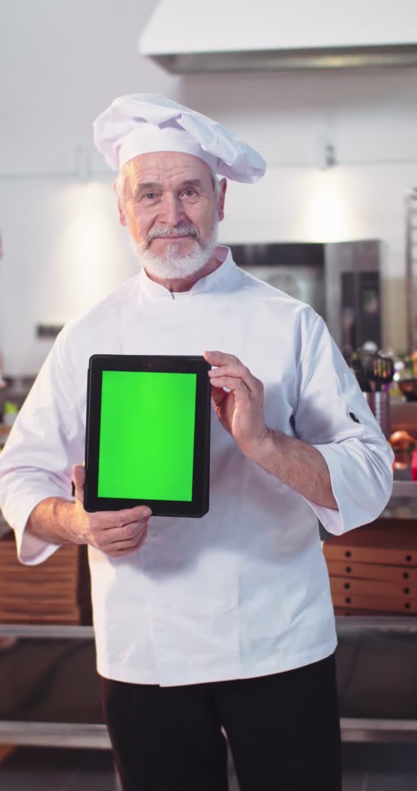 Portrét šťastného kavkazského starého kuchaře stojícího v kuchyni v uniformě, držícího tabletu se zelenou obrazovkou a hledícího do kamery. Vertikální záběry Radostný senior kuchař v restauraci, chroma klíč — Stock video