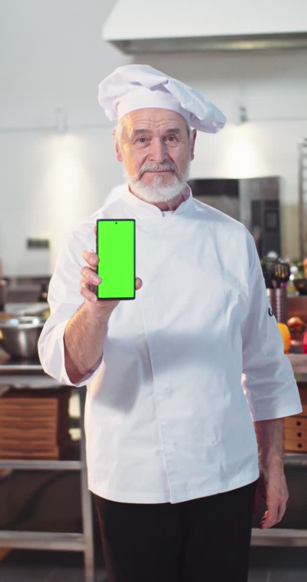 Retrato de caucásico feliz hombre mayor cocinero de pie en la cocina en uniforme mostrando teléfono inteligente con pantalla verde y mirando a la cámara. Vídeo vertical. Anciano chef masculino en restaurante propio, croma key — Vídeos de Stock