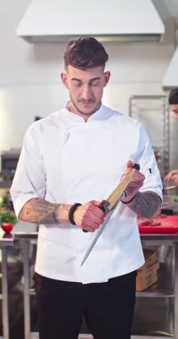 Kafkasyalı genç yakışıklı, dövmeli profesyonel aşçının dikey videosu. Lokanta mutfağında duruyor. Bıçağı biliyor, yemek pişirmeye ve kameraya bakıyor. Restoran, aşçılık konsepti.