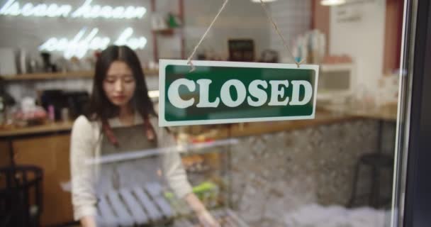 Joven propietaria de una cafetería que se acerca a la ventana de la tienda y mira afuera. Mujer de negocios asiática abriendo café después del cierre, poniendo anuncio Mantenga la distancia social, y volteando signo CERRADO. — Vídeo de stock