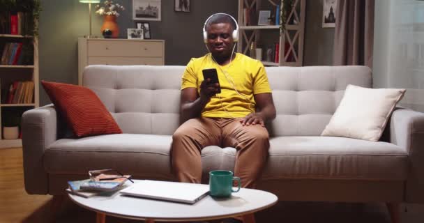 Dobře vypadající Afroameričan sedí na gauči a doma píše kamarádovi na smartphonu. Mladý muž ve sluchátkách poslouchá hudbu, relaxuje a baví se večer. Zábavní koncept. — Stock video